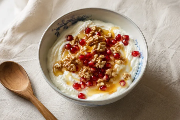 A bowl of Greek yogurt topped with honey, walnuts, and pomegranate seeds on a bone-linen surface